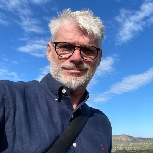 Torsten Forberg Homeservice Hausbetreuung Mallorca Mann mit grauem Haar und Brille, trägt ein dunkles Hemd, lächelt vor blauem Himmel. Torsten Forberg Homeservice Mallorca