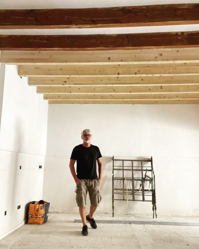 Baubegleitung durch Torsten Mann in schwarzem Shirt und kurzen Hosen in einem leeren Raum mit Holzdeckenbalken.