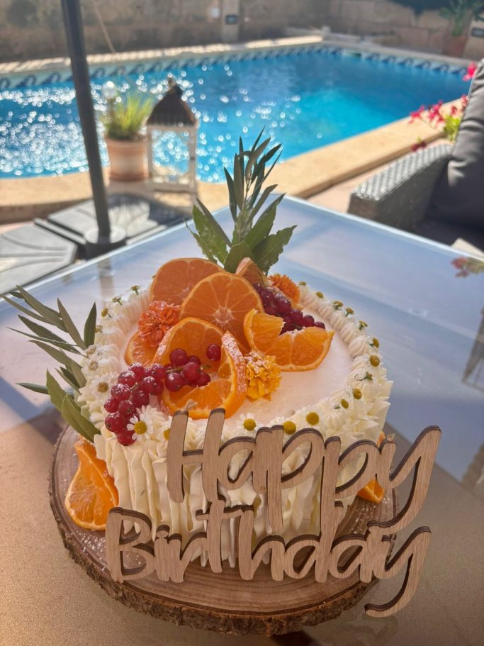 Geburtstagstorte Geburtstagskuchen mit Früchten und einem Holzschild „Happy Birthday“ vor einem Pool.