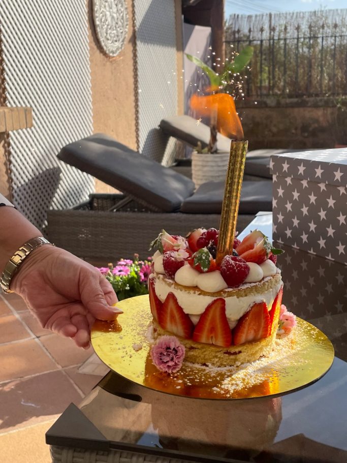 Dulce de Anja - Mallorca Erdbeerkuchen mit Sahne, dekoriert mit Früchten und einer Kerze, auf einem goldenen Teller.