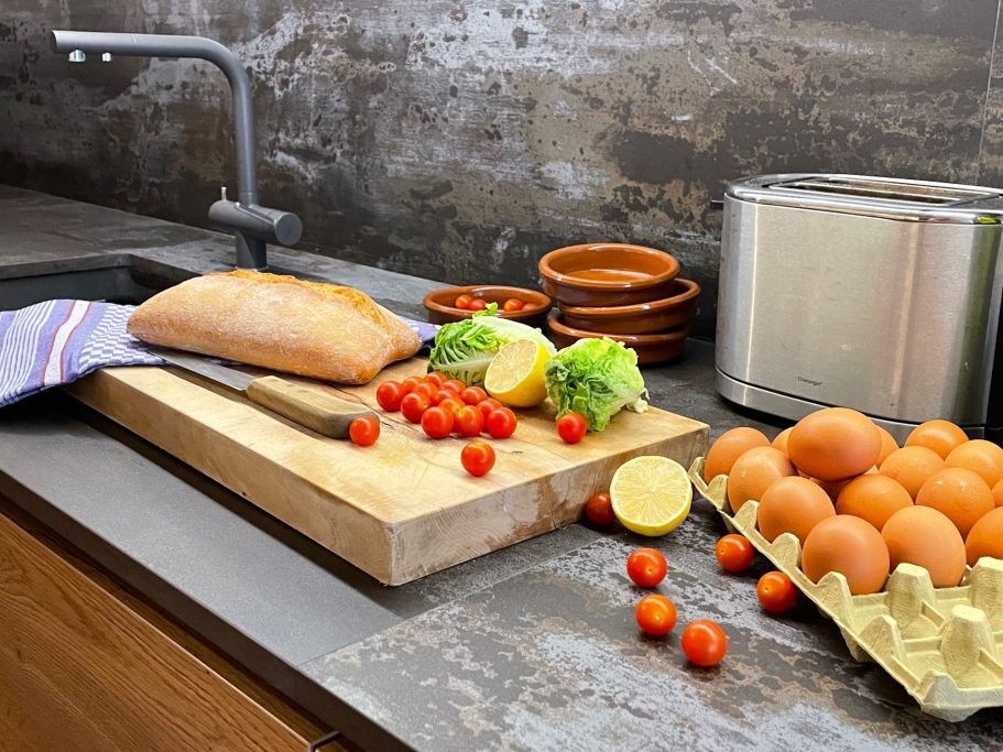 Gästebetreuung Brett mit frischem Brot, Tomaten, Eiern, Salat und Zitronen in einer modernen Küche.