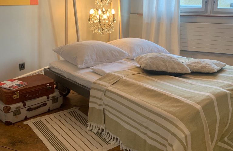 Gemütliches Schlafzimmer mit Bett, Kissen, Teppich und einem alten Reisekoffer. Ferienvermietung Mallorca