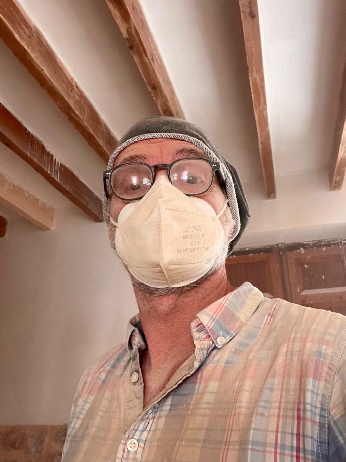 Torsten bei Schleifarbeiten Mann mit Brille und Beanie trägt eine helle Maske in einem Zimmer mit Holzbalken.