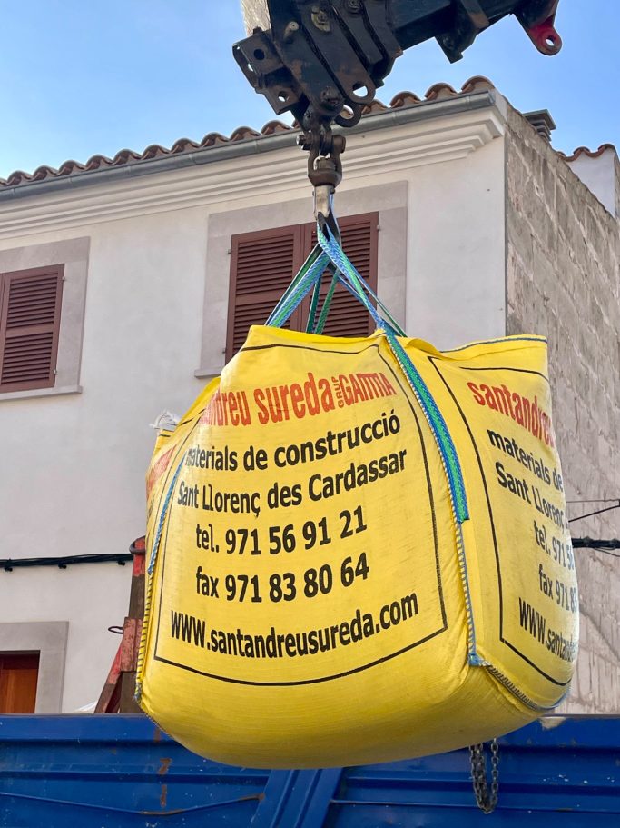 Baubegleitung Gelbe Baugrube-Tasche wird von einem Kran angehoben, im Hintergrund ein Gebäude.