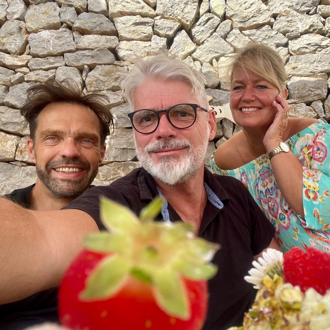 Ein Team von erfahrenen und serviceorientierten langjährigen Freunden, die sich für Ihre Wünsche um Ihre Immobilie einsetzen. Anja Gerd und Torsten Drei Personen mit Blumen vor einer steinernen Wand. Team im Homeservice - Anja, Gerd und Torsten